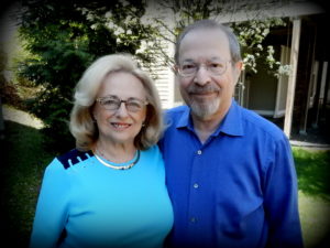 woman with shoulder-length light blond hair and man with goatee, both wearing glasses, in front of a house