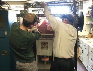 two men work on a machine with welding shields and thick gloves