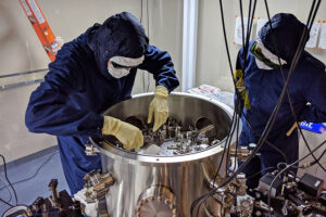 Nicholas Demos and another scientist in lab suits, gloves, and googles before a metal drum