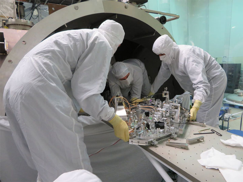 Three people in hazmat suits lift a silver piece of equipment with wires