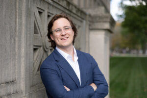 Davis Evans stands in a blue blazer and white shirt, smiling and leaning against a wall.