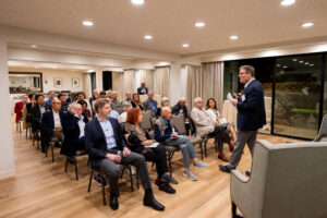 Michale Fee stands at the front of a room addressing a seated crowd